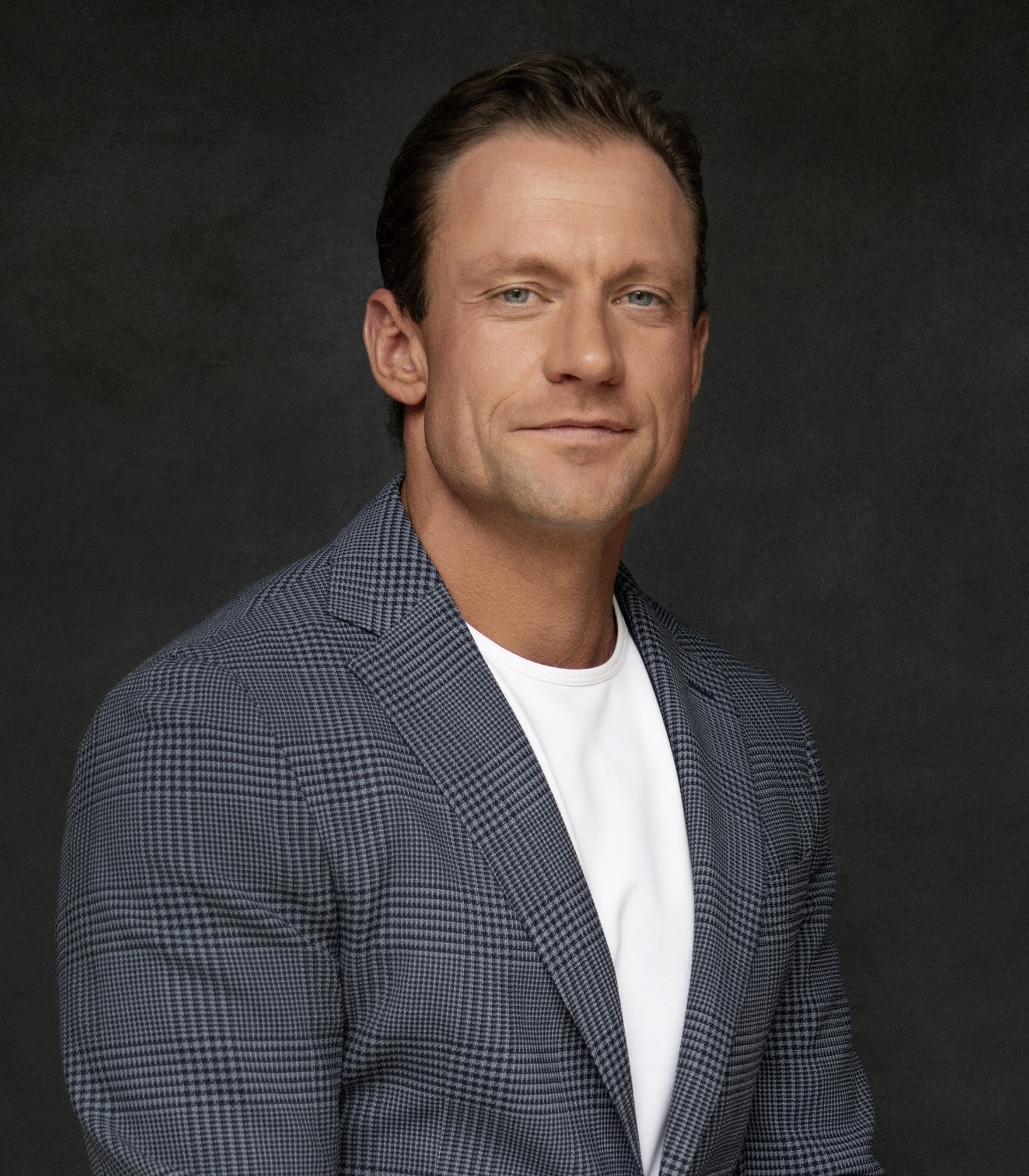 Portrait of a man wearing a blue checkered blazer over a white T-shirt against a dark backdrop, looking at the camera with a slight smile.