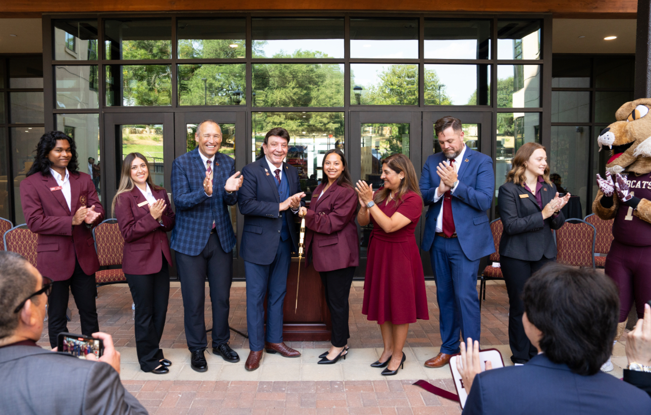 Group of staff in burgundy blazers applaud in front of a glass-door entrance, with a lion mascot on the right and spectators seated nearby.