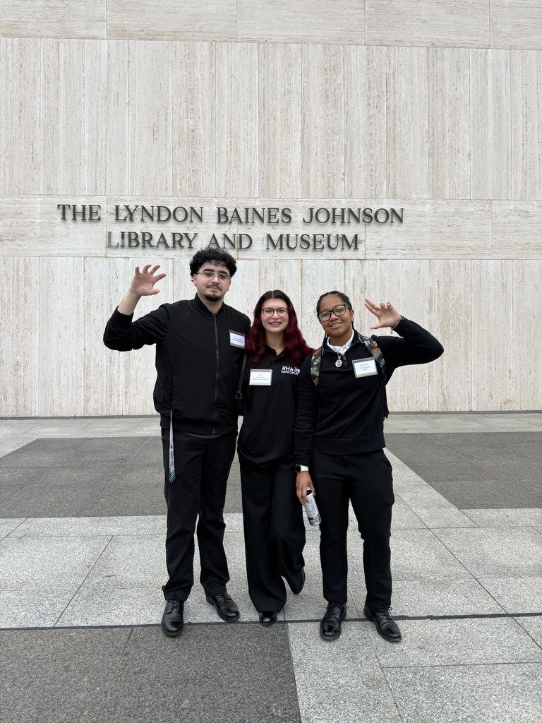 Three people in black outfits pose and wave in front of the Lyndon Baines Johnson Library and Museum sign on a stone wall.