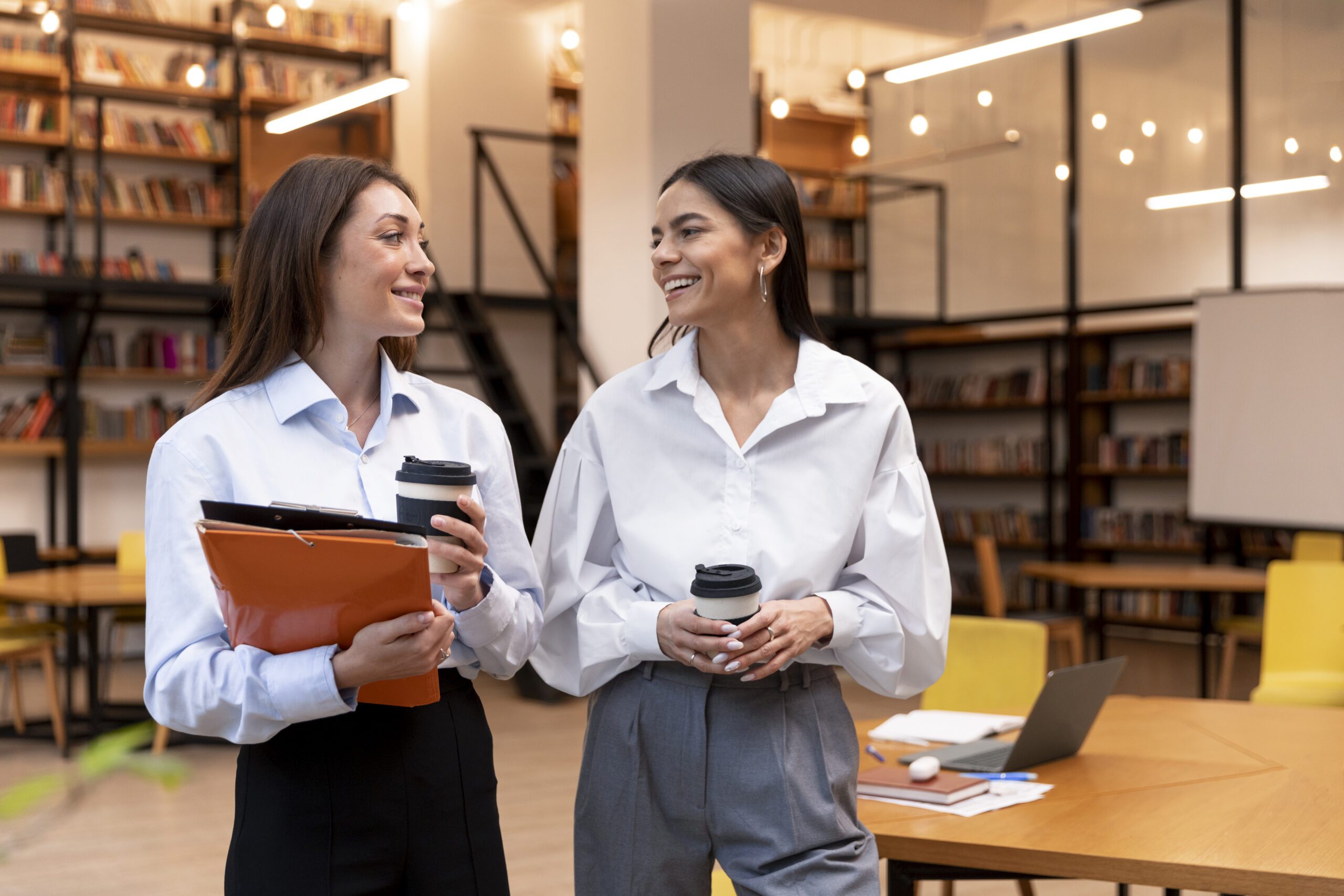 Two women in a library lounge smile at each other, holding coffee cups and folders.