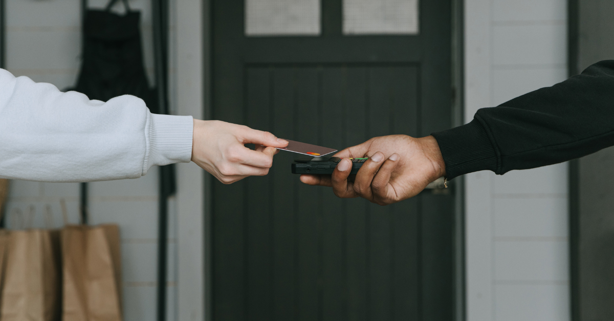 Person in a white sleeve hands a card to another person holding a card reader at a doorway—suggesting a payment or authentication action.