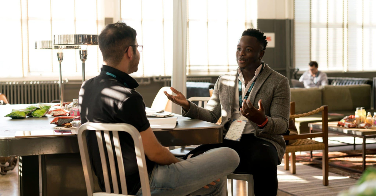 Two men seated at a table in a bright lounge, engaged in conversation; one wears a gray blazer and conference badge while the other faces away.